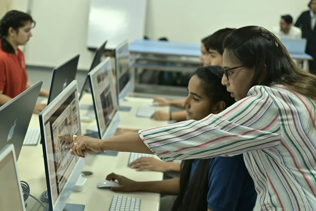 An instructor helping learners at desktop computers in an illustrative training room.